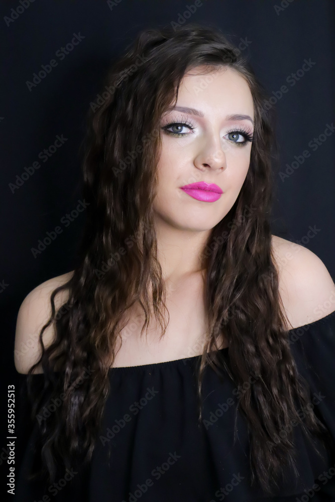 beautiful brunette young girl   looking at camera with fresh make up on black background, pink lipstick curly hair over shoulders, in black dress  studio shot, vertical image