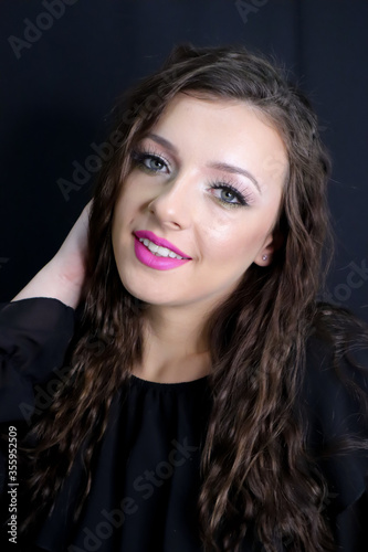 beautiful brunette young girl happily smiling, looking at camera with fresh make up on black background, pink lipstick curly hair over shoulders, in black dress studio shot, vertical image