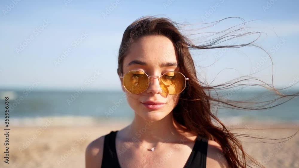 Portrait of young woman looking at camera standing on the beach with a sea on background. Attractive girl in black swimsuit and sunglasses raises eyes. Wind blows airy brown hair shining in sun. Portrait of young woman looking at camera standing on the beach with a sea on background. Attractive girl in black swimsuit and sunglasses raises eyes. Wind blows airy brown hair shining in sun.