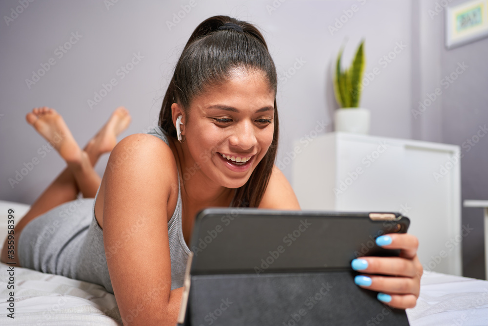 Naklejka premium Young Latina studying and performing class assignments online with his computer from home for causes of coronavirus. Sitting at her table with leaves and pens. Videoconferencia con sus profesores.