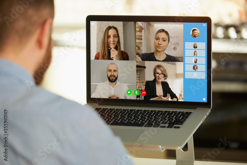 Back view of a man who working remotely and listening to colleagues about business in a video call on a laptop at home. A close photo of a computer display with a business team at an online meeting.