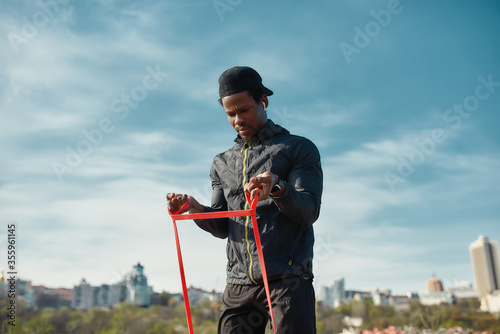 Working out in the morning. Young strong african man in sportswear exercising, using resistance band outdoors against blurred city background