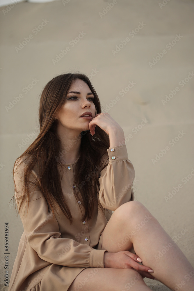 Young beautiful girl model thinking and sitting on the beach in dress. Nature sand background Young beautiful girl model thinking and sitting on the beach in dress. Nature sand background