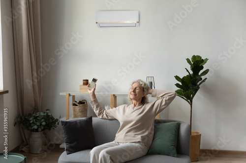 Happy senior 50s woman relax sit on cozy couch in living room turn on modern conditioner with remoter controller, smiling elderly grandmother breathe enjoy fresh air under AC gadget at home photo