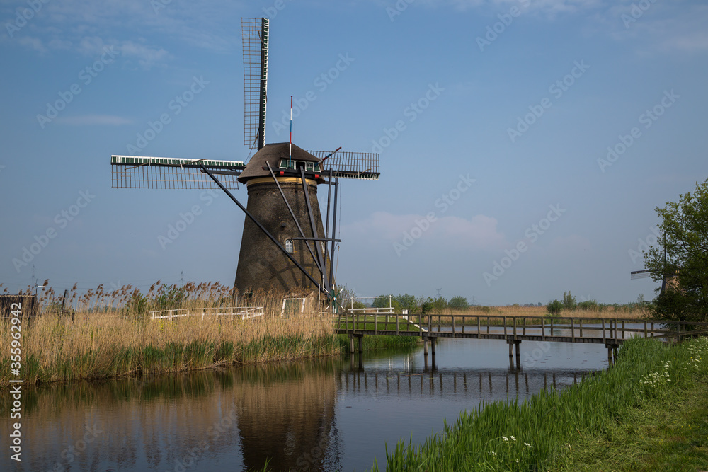 Historic windmill at Kinderdijk, Netherlands hemmed in with reeds at the side of a canal crossed by a wooden foot bridge.