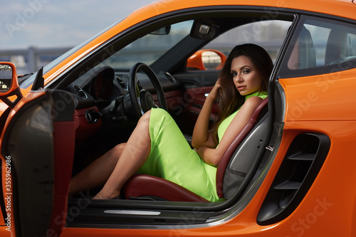 Young beautiful brunette woman in green neon dress sitting in orange sport car. Fashion shot