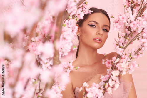 Closeup fashion spring face portrait of young beautiful caucasian woman with brunette hair in pony tail and perfect makeup looking through trees with pink flowers in blossom