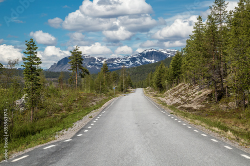 BD 805, a narrow, lonely road leading through the arctic wilderness of Lapland, Sweden, connecting Jokkmokk and Kvikkjokk. A rugged mountain range covered with snow is visible in the background.