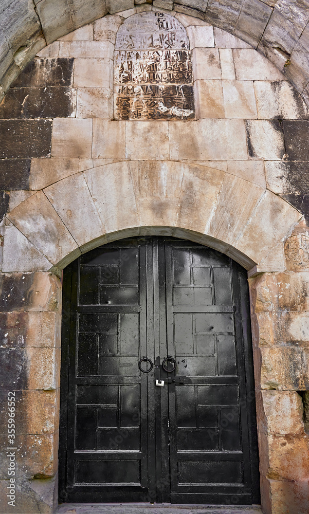 Black cast iron forged door, selective focus. Ancient antique ancient door in the arch and the ancient inscriptions above it. Textured stone tiles.