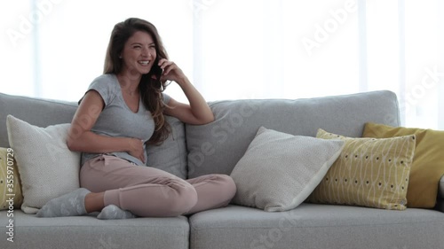 Young beautiful smiling girl sitting on the sofa in her living room talking and chit chat on the phone with her girlfriend about boys and laughing in casual clothes 