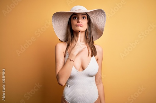 Young beautiful brunette woman on vacation wearing swimsuit and summer hat Thinking concentrated about doubt with finger on chin and looking up wondering