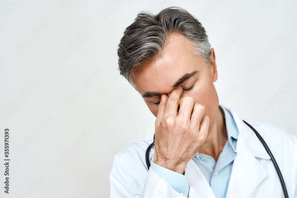 Stress at work. Close up portrait of tired unhappy male doctor feeling upset, keeping eyes closed and touching nose while standing against grey background Stress at work. Close up portrait of tired unhappy male doctor feeling upset, keeping eyes closed and touching nose while standing against grey background