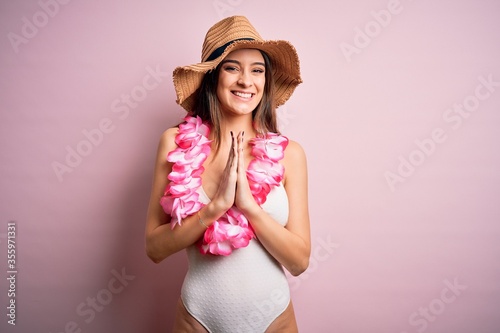 Young beautiful brunette woman on vacation wearing swimsuit and Hawaiian flowers lei praying with hands together asking for forgiveness smiling confident.