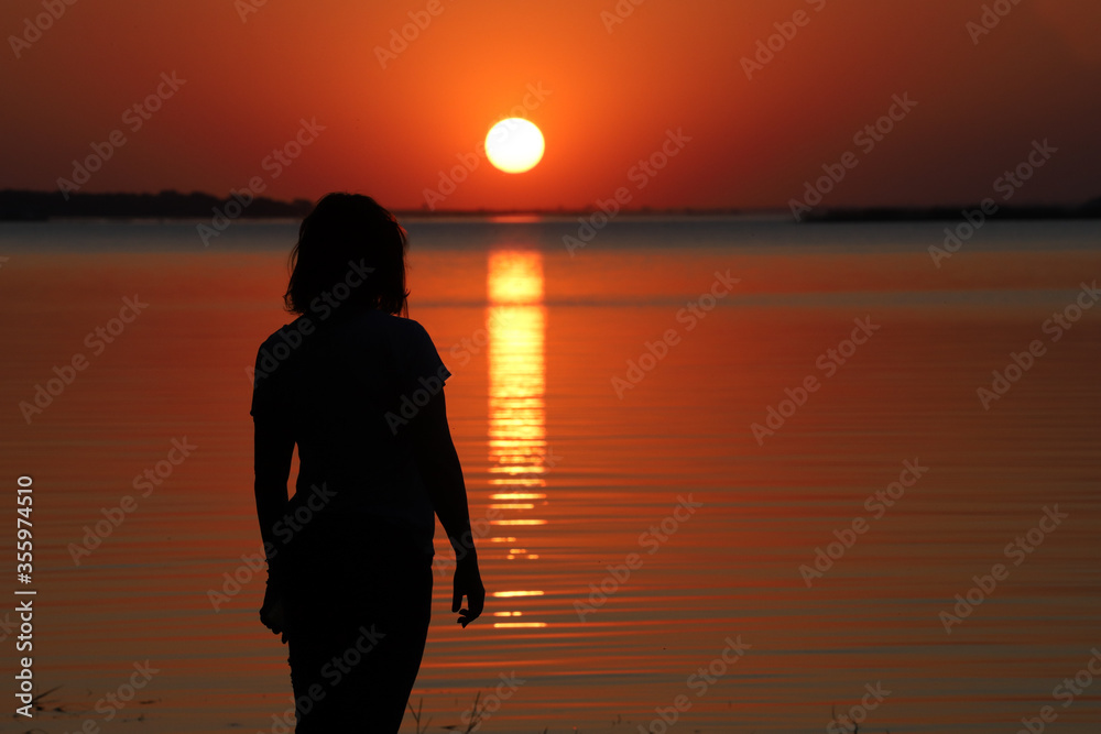 silhouette of woman admiring the sunset in golden, yellow and red tones, with the sun descending over the waters of a river.