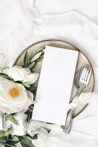 Festive summer wedding scene. Marble table setting with cutlery, olive branches, white peony flowers, stoneware plate and silk ribbon. Blank restaurant menu card mockup. Flat lay, top view, vertical.