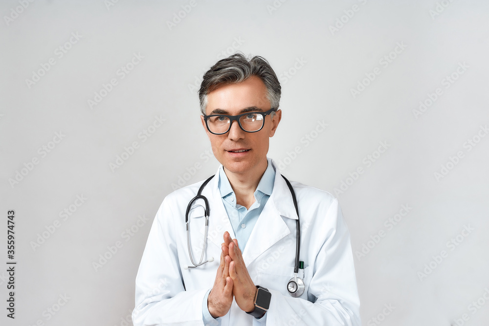 Portrait of mature male doctor or physician in white coat with stethoscope around neck praying to God for help while standing against grey background Portrait of mature male doctor or physician in white coat with stethoscope around neck praying to God for help while standing against grey background