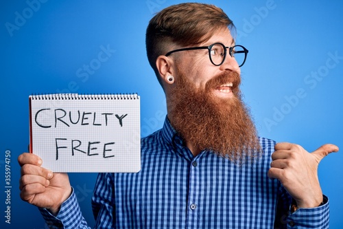Redhead Irish man with beard holding cruelty free vegan cosmetics message over blue background pointing and showing with thumb up to the side with happy face smiling