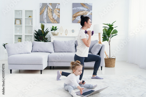 Funny baby girl playing with laptop while her sporty healthy mommy having online yoga training at home.