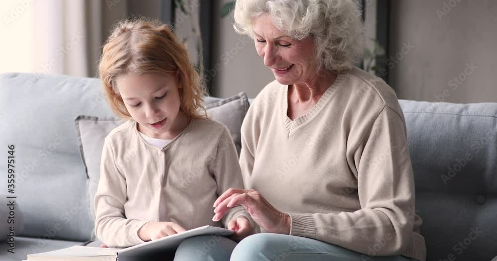 Cute little redhead child girl learning reading paper book with happy middle aged grandmother, sitting together on couch indoors. Smiling senior old woman feeling proud of smart small granddaughter. Cute little redhead child girl learning reading paper book with happy middle aged grandmother, sitting together on couch indoors. Smiling senior old woman feeling proud of smart small granddaughter.