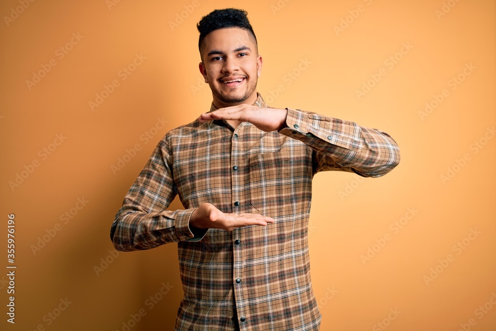 Young handsome man wearing casual shirt standing over isolated yellow background gesturing with hands showing big and large size sign, measure symbol. Smiling looking at the camera. Measuring concept. Young handsome man wearing casual shirt standing over isolated yellow background gesturing with hands showing big and large size sign, measure symbol. Smiling looking at the camera. Measuring concept.