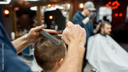 Cropped photo of barber with scissors and hair comb in hands making a haircut for little boy in the modern barbershop. Selective focus