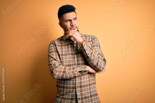 Young handsome man wearing casual shirt standing over isolated yellow background with hand on chin thinking about question, pensive expression. Smiling with thoughtful face. Doubt concept.