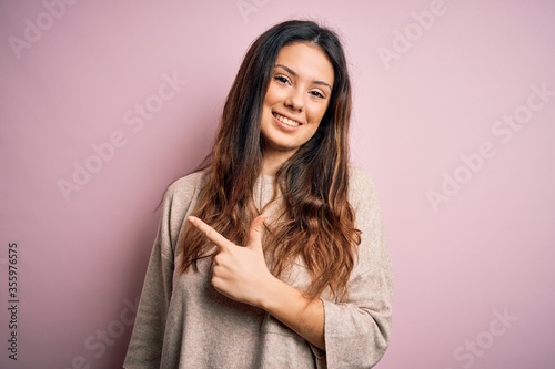 Young beautiful brunette woman wearing casual sweater standing over pink background cheerful with a smile on face pointing with hand and finger up to the side with happy and natural expression