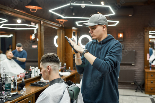 Working in barbershop. Professional barber in a baseball cap doing hair styling for young and handsome guy sitting in chair in the front of a mirror
