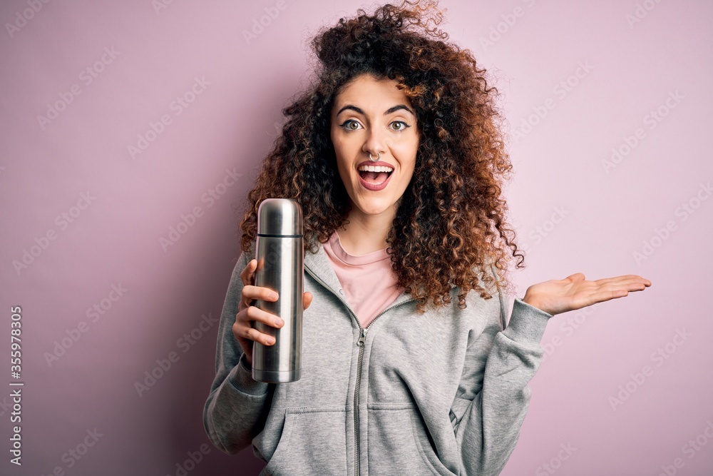 Young beautiful tourist woman with curly hair and piercing holding thermo with water very happy and excited, winner expression celebrating victory screaming with big smile and raised hands Young beautiful tourist woman with curly hair and piercing holding thermo with water very happy and excited, winner expression celebrating victory screaming with big smile and raised hands