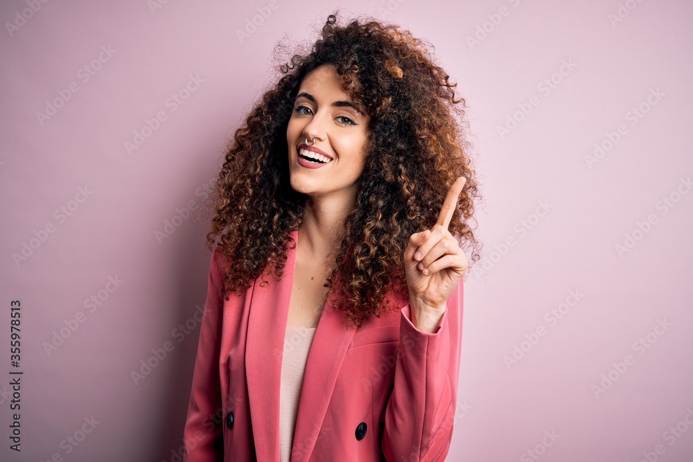 Young beautiful businesswoman with curly hair and piercing wearing elegant jacket pointing finger up with successful idea. Exited and happy. Number one. Young beautiful businesswoman with curly hair and piercing wearing elegant jacket pointing finger up with successful idea. Exited and happy. Number one.