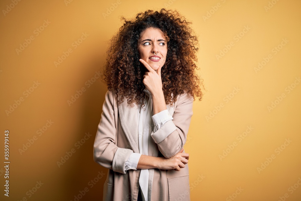 Young beautiful brunette woman with curly hair and piercing wearing casual t-shirt and diadem with hand on chin thinking about question, pensive expression. Smiling with thoughtful face. Doubt