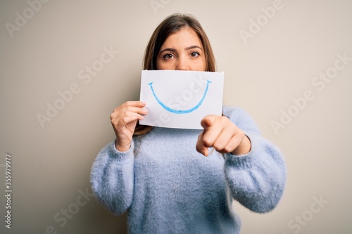 Young blonde woman holding funny smile drawing on mouth as happy expression pointing with finger to the camera and to you, hand sign, positive and confident gesture from the front