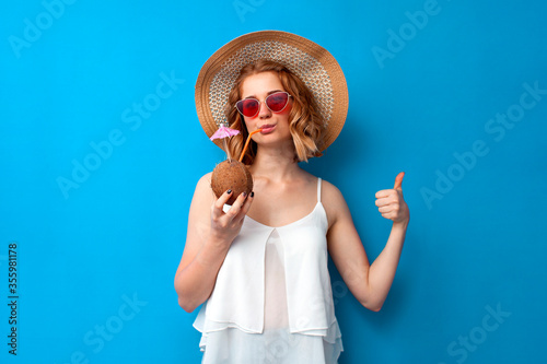 girl in summer clothes drinks a coconut cocktail and shows like on a blue isolated background, woman tourist in a summer resort