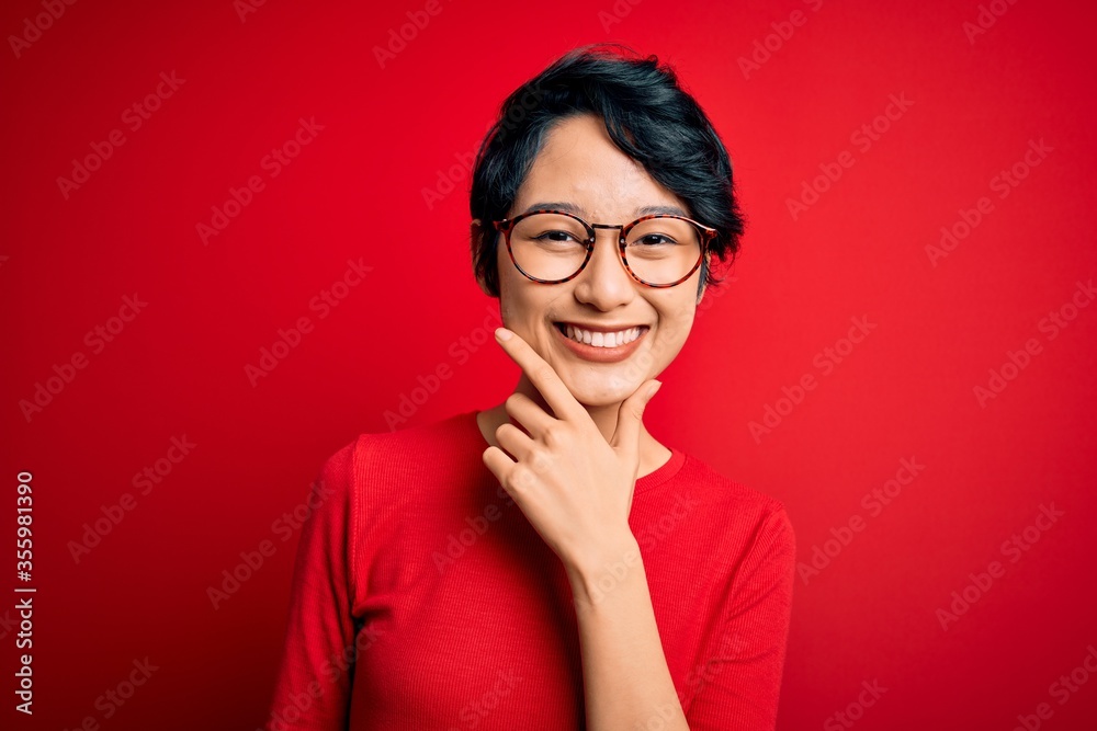 Young beautiful asian girl wearing casual t-shirt and glasses over isolated red background looking confident at the camera smiling with crossed arms and hand raised on chin. Thinking positive.