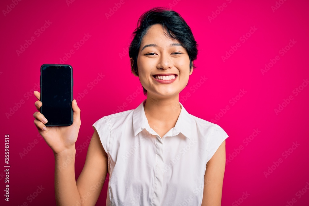 Young beautiful asian girl holding smartphone showing screen over isolated pink background with a happy face standing and smiling with a confident smile showing teeth Young beautiful asian girl holding smartphone showing screen over isolated pink background with a happy face standing and smiling with a confident smile showing teeth