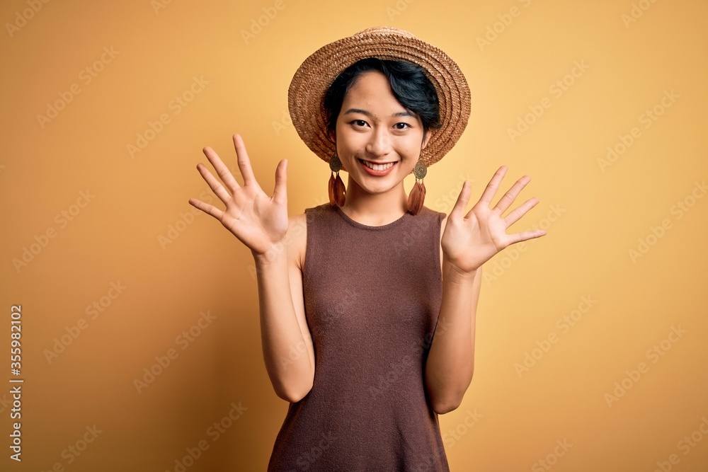 Young beautiful asian girl wearing casual t-shirt and hat over isolated yellow background showing and pointing up with fingers number ten while smiling confident and happy. Young beautiful asian girl wearing casual t-shirt and hat over isolated yellow background showing and pointing up with fingers number ten while smiling confident and happy.