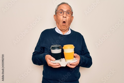 Senior handsome hoary man holding tray with takeaway cups of coffee over white background scared in shock with a surprise face, afraid and excited with fear expression