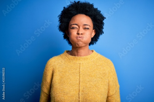 Young beautiful African American afro woman with curly hair wearing yellow casual sweater puffing cheeks with funny face. Mouth inflated with air, crazy expression.