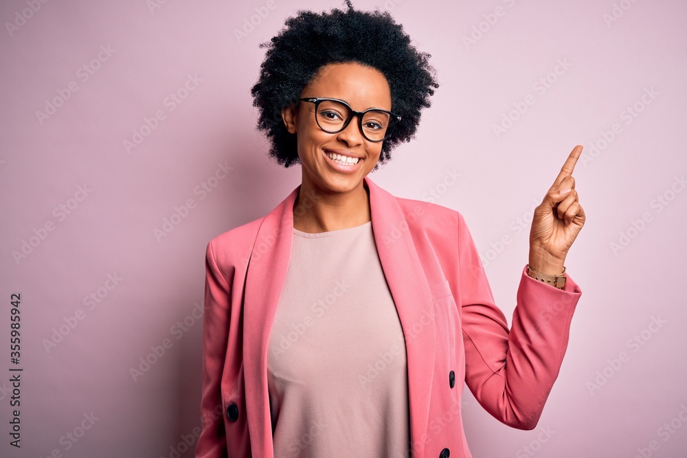 Young beautiful African American afro businesswoman with curly hair wearing pink jacket with a big smile on face, pointing with hand finger to the side looking at the camera. Young beautiful African American afro businesswoman with curly hair wearing pink jacket with a big smile on face, pointing with hand finger to the side looking at the camera.