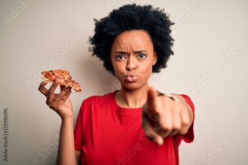 Young African American afro woman with curly hair eating slice of delicious Italian pizza pointing with finger to the camera and to you, hand sign, positive and confident gesture from the front