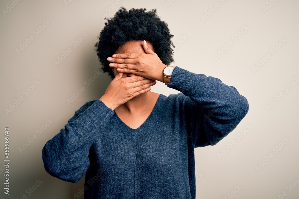 Young beautiful African American afro woman with curly hair wearing casual sweater Covering eyes and mouth with hands, surprised and shocked. Hiding emotion Young beautiful African American afro woman with curly hair wearing casual sweater Covering eyes and mouth with hands, surprised and shocked. Hiding emotion
