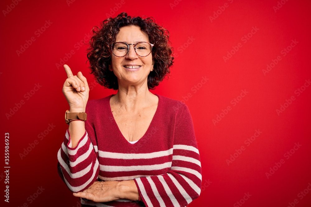 Middle age beautiful curly hair woman wearing casual striped sweater over red background with a big smile on face, pointing with hand and finger to the side looking at the camera. Middle age beautiful curly hair woman wearing casual striped sweater over red background with a big smile on face, pointing with hand and finger to the side looking at the camera.