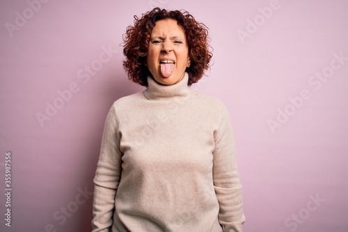 Middle age beautiful curly hair woman wearing casual turtleneck sweater over pink background sticking tongue out happy with funny expression. Emotion concept.