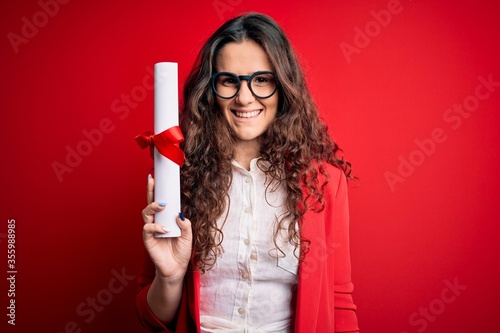 Young beautiful woman with curly hair holding university diploma degree over red background with a happy face standing and smiling with a confident smile showing teeth photo