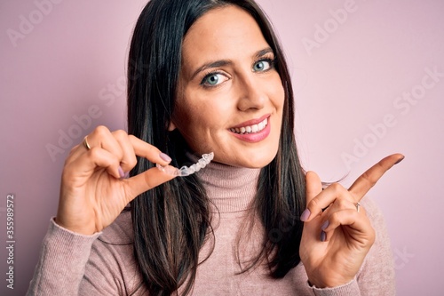 Young woman with blue eyes holding clear aligner standing over pink background very happy pointing with hand and finger to the side