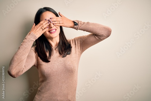 Young brunette woman with blue eyes wearing casual sweater over isolated white background covering eyes with hands smiling cheerful and funny. Blind concept.
