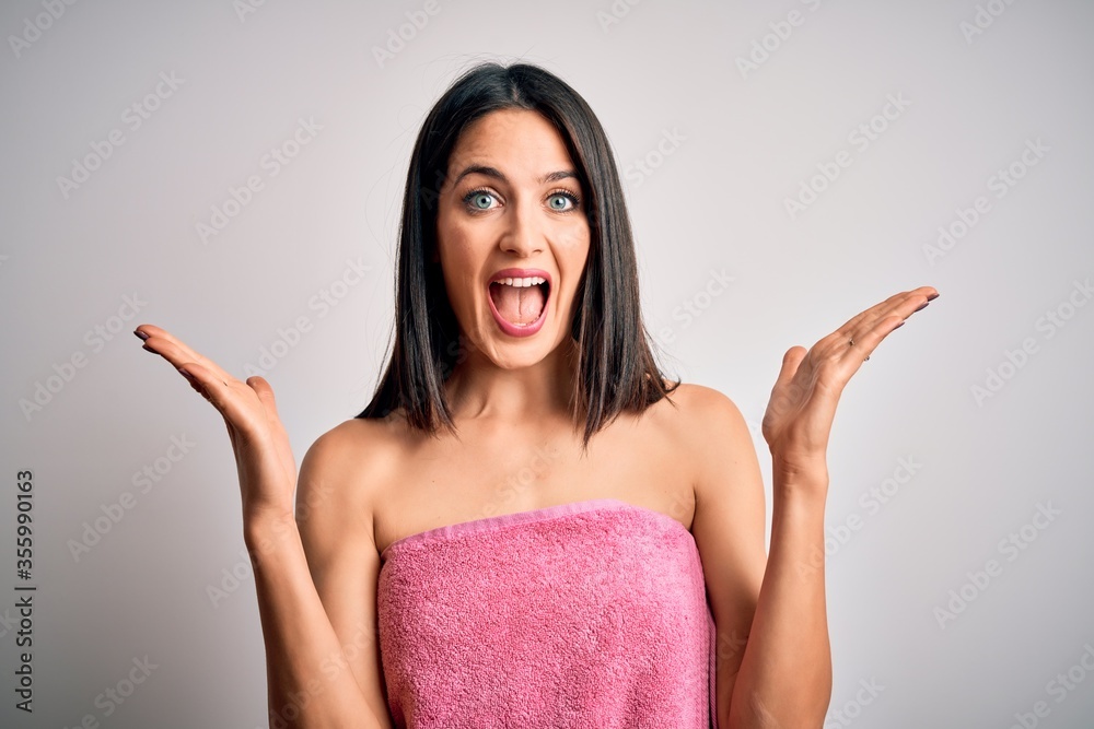 Young brunette woman with blue eyes wearing shower towel after bath over white background celebrating crazy and amazed for success with arms raised and open eyes screaming excited. Winner concept Young brunette woman with blue eyes wearing shower towel after bath over white background celebrating crazy and amazed for success with arms raised and open eyes screaming excited. Winner concept