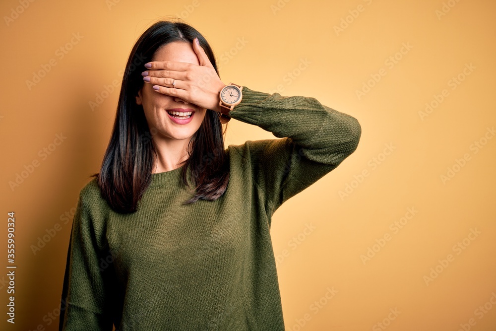 Φωτογραφία Young brunette woman with blue eyes wearing green casual sweater over yellow background smiling and laughing with hand on face covering eyes for surprise
