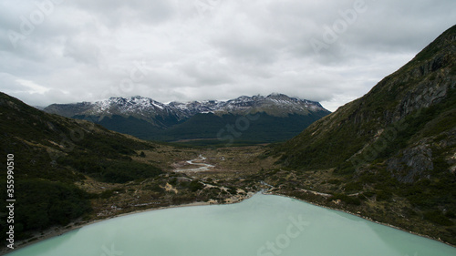 Aerial view of glacier water Emerald Lake in the mountaintop in Ushuaia, Tierra del Fuego, Patagonia Argentina. A stream flows downhill the forest and valley into the Andes mountains with snowy peaks