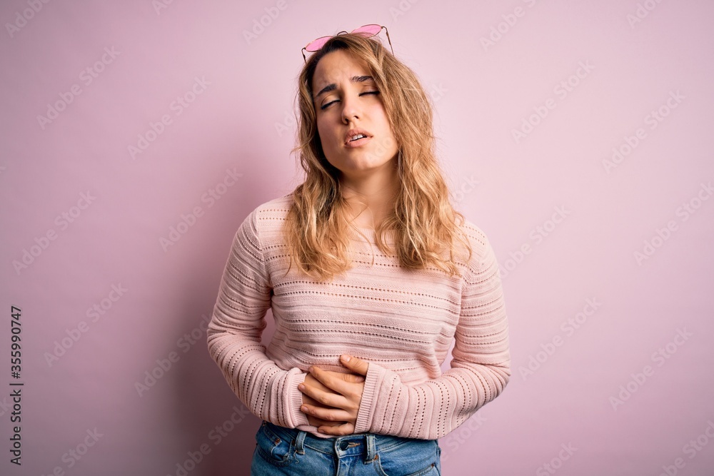 Young beautiful blonde woman wearing casual sweater and sunglasses over pink background with hand on stomach because indigestion, painful illness feeling unwell. Ache concept. Young beautiful blonde woman wearing casual sweater and sunglasses over pink background with hand on stomach because indigestion, painful illness feeling unwell. Ache concept.